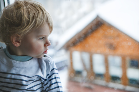 Little toddler boy looking out of the window on winter day with snow landscapeの写真素材