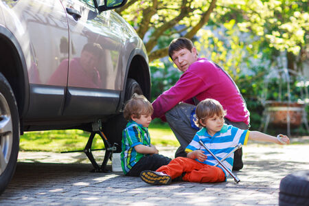 Happy family of three: father and two little boys repairing car and changing wheel together on warm day, outdoors.の写真素材