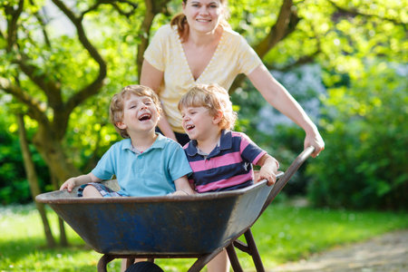 Two little boys having fun in a wheelbarrow pushing by mother in summer gardenの写真素材