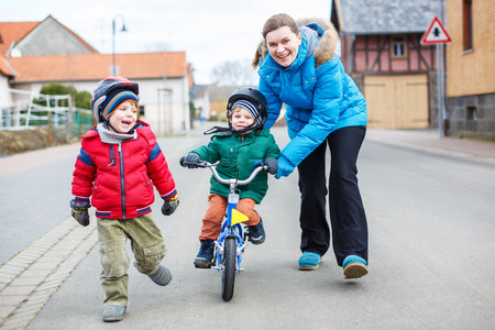 Young mother teaching her 2 years old little son to ride a bike, outdoors. Another sibling child running beside.の写真素材
