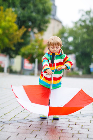Happy little boy walking in city and playing with red umbrella, wearing colorful rain coat and green boots outdoors at rainy dayの写真素材