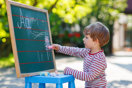Cheerful boy at blackboard practicing writing letters, outdoor school or nursery