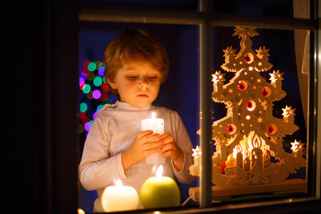 Little boy standing by window at Christmas time and holding candle. With colorful lights from Christmas tree on background, selective focus.の写真素材