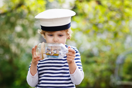 Happy kid boy in skipper uniform playing with toy ship against green tree summer backgroundの写真素材