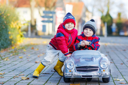 Two happy sibling boys in red jackets and rain boots playing with big old toy car, outdoors.  Kids leisure on cold day in winter, autumn or spring.の写真素材