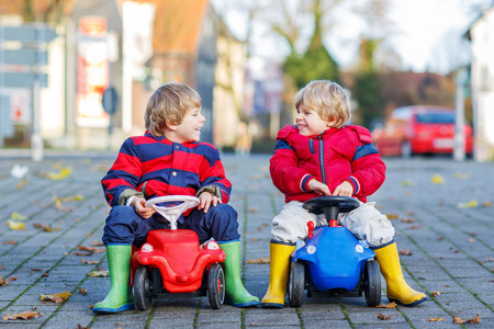 Two little twinds and friends boys in red jackets and rain boots playing with colorful toy cars, outdoors. Kids leisure on cold day in winter, autumn or spring.の写真素材