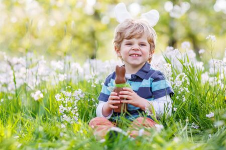 Happy little toddler boy eating chocolate and wearing Easter bunny ears, sitting in blooming garden on warm sunny day. Celebrating Easter traditional holiday.の写真素材