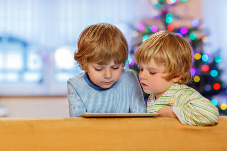 Two happy little kids playing with tablet pc, indoors. Sibling toddlers looking at tablet. With Christmas tree and lights on background.の写真素材