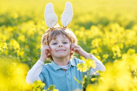 Funny blond kid of 3 years with Easter bunny ears playing yellow rape field on sunny spring day, celebrating Easter holidayの写真素材
