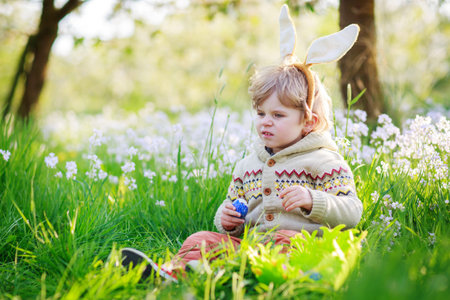 Cute happy little boy wearing Easter bunny ears at spring green grass and blooming apple garden,...