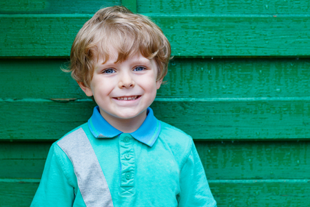 Portrait of happy little cute kid boy with blond hairs and blue eyes against green wooden background in gardenの写真素材
