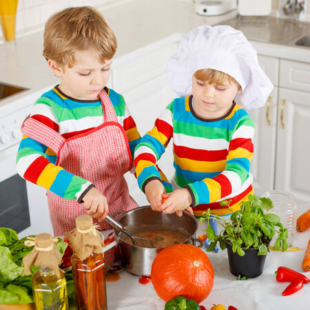 Little twins cooking italian soup and meal with fresh vegetables in home's white kitchen. Sibling children in colorful shirts.の写真素材