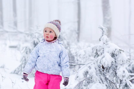 portrait of a little girl in winter hat in snow forest at snowflakes background. outdoors winter leisure and lifestyle with kids.の写真素材
