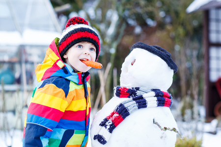 Funny little kid boy making a snowman and eating carrot, playing and having fun with snow, outdoors  on cold day. Active outoors leisure with children in winter.の写真素材