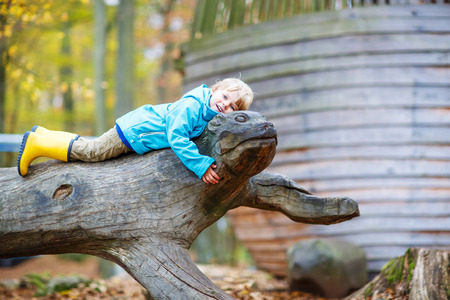 Cute little toddler boy in blue rain jacket and gumboots having fun with playing on playground on warm, autumn day, outdoorsの写真素材