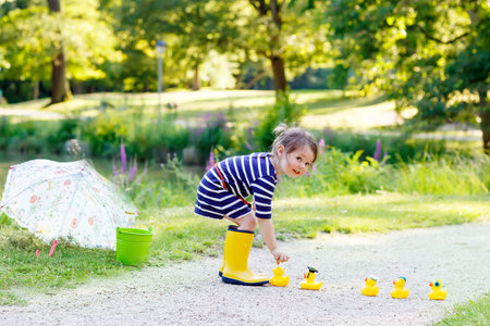 Cute little kid girl in yellow rainboots playing with yellow rubber ducks in summer park. Creative leisure with kids in forest on sunny day.の写真素材