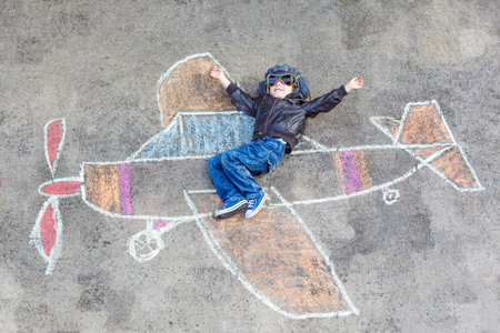 Happy little kid boy in pilot uniform having fun with airplane picture drawing with colorful chalk. Creative leisure for children outdoors in summer.の写真素材