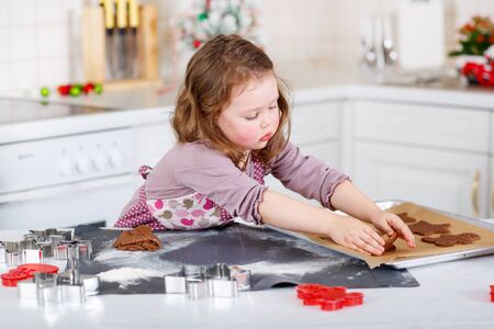 Happy little child, cute kid girl, sitting at the table in domestic kitchen making delicious sweet gingerbread xmas cookies. Kitchen decorated for Christmasの写真素材