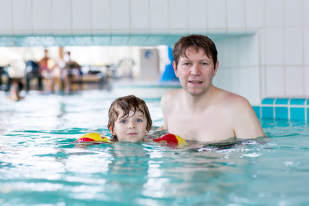 Cute boy learning swimming with dad in indoor pool. Active happy kid boy wearing safe swimmies....