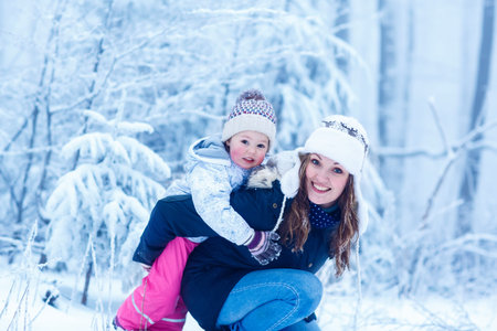 Happy young mum and her little daughter in winter hats in snow forest at snowflakes . outdoors winter leisure and lifestyle with kids.の写真素材