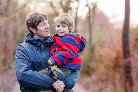 Happy dad holding his son, little funny kid boy on arm, on cold day, walking together in a park in...