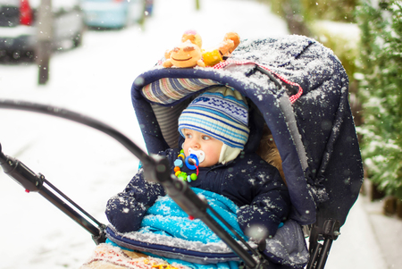 Cute baby boy in warm clothes in pram during winter snow fall on cold winter day. Happy carefree childhood.の写真素材