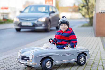 Funny cute kid boy in red bright jacket driving big vintage old toy car and having fun, outdoors. Active leisure for kids on cold day in winter, autumn or spring.の写真素材