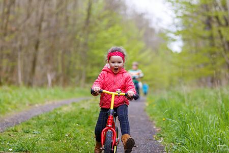 Happy funny kid girl and colorful raincoat riding his first bike on summer day. Active leisure for children outdoors.の写真素材