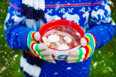 Hands of child holding big cup with snowflakes and hot cocoa and chocolate drink and marshmallows. Kid in winter sweater, long warm scarf and colorful gloves. On cold winter day.の写真素材