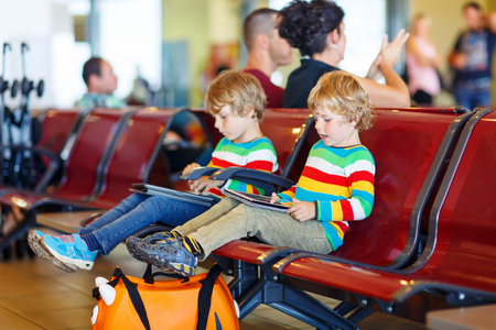 Two tired little sibling kids boys at the airport, traveling together. Upset children waiting and playing with tablet pc. Canceled flight due to pilot strike.の写真素材