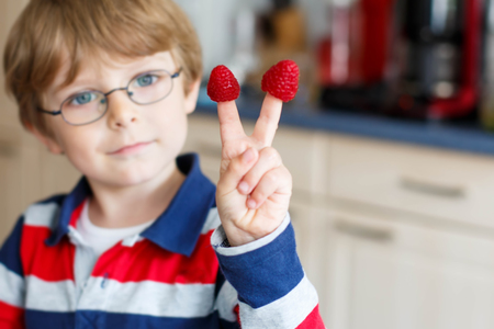 Happy adorable kid boy with glasses eating healthy food in kindergarten or at home. Fresh raspberries as snack for children. Selective focus on fingersの写真素材