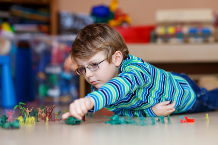 Cute blond child playing with lots of small toy soldiers, indoor. Active kid boy with glasses wearing colorful shirt and having fun at home or at nursery.の写真素材