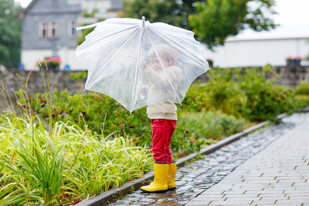 Little blond kid boy walking with big umbrella outdoors on rainy day. Child having fun and wearing colorful waterproof clothes and rain boots.の写真素材