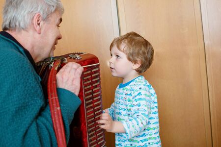 Happy blond little kid boy and his grandfather playing together with accordion. Senior man teaching his grandson, cute toddler to play with music instrument at home.の写真素材
