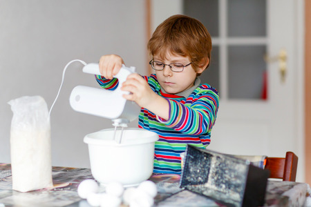 Active funny blond little kid boy n glasses baking apple cake and muffins in domestic kitchen. Happy child having fun with working with mixer, flour, eggs and fruits at home.の写真素材