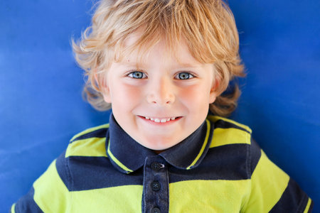 Portrait of cute happy blond little kid boy against blue background. child smiling and looking at...