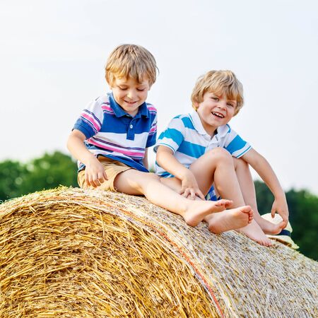Two little children and friends sitting on hay stack or bale and speaking on yellow wheat field in summer. Active outdoors leisure with children on warm summer day.の写真素材