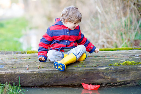 Beautiful little kid boy in rain boots playing with paper boat by a puddle on warm spring day. Active leisure for children. Child having fun outdoors.の写真素材