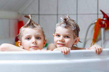 Happy siblings: Two little twins children playing together with water by taking bath in bathtub at home. Kid boys having fun together.の写真素材