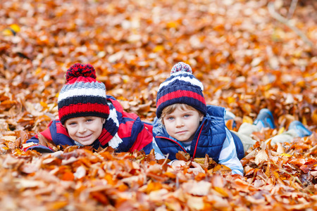 Two little brothers boys lying in autumn leaves in colorful clothing. Happy siblings kids having fun in autumn forest or park on warm fall day. With hats and scarfsの写真素材