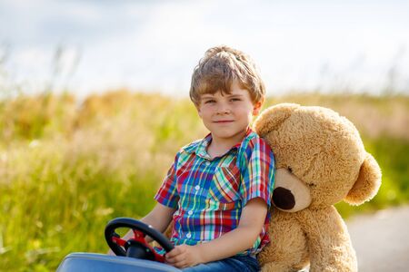 Little preschool kid boy driving big toy car and having fun with playing with his plush toy bear, outdoors. Child enjoying warm summer day in nature landscapeの写真素材