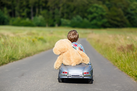 Little preschool kid boy driving big toy car and having fun with playing with his plush toy bear, outdoors. Child enjoying warm summer day in nature landscapeの写真素材