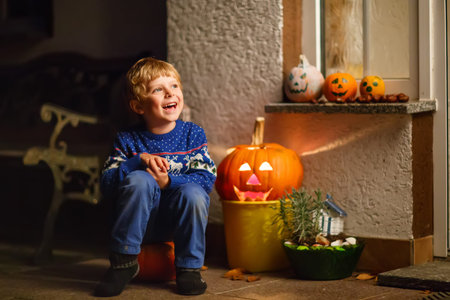 Little kid boy sitting with traditional jack-o-lanterns pumpkins for halloween by the decorated scary door, outdoors. Child having fun and celebrating holiday.の写真素材