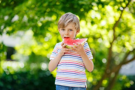 Adorable little preschool kid boy with blond hairs eating watermelon in summer garden. Funny happy child smiling and tasting healthy fruit snack on sunny day.の写真素材