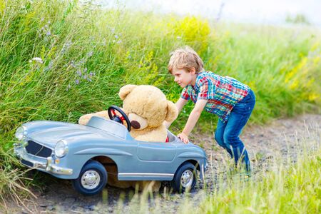 Little preschool kid boy driving big toy car and having fun with playing with his plush toy bear, outdoors. Child enjoying warm summer day in nature landscapeの写真素材
