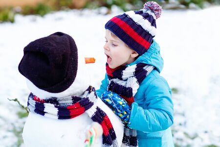 Funny little kid boy making a snowman and eating carrot, playing and having fun with snow, outdoors  on cold day. Active leisure with children in winter.の写真素材