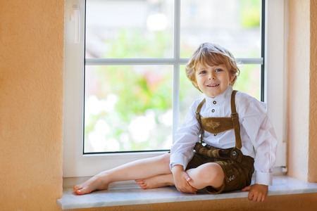 Adorable little blond kid boy sitting near window and looking on raindrops on cold spring or autumn day, indoors. Happy child.の写真素材