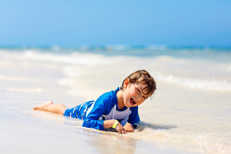 Adorable little blond kid boy having fun on tropical beach of Maldives. Excited child playing and surfing in sun protected swimsuit in ocean on vacations.の写真素材