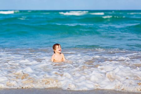 Adorable little blond kid boy having fun on ocean beach. Excited child playing with waves, swimming and happy about family vacations in Miami, Florida, USA.の写真素材