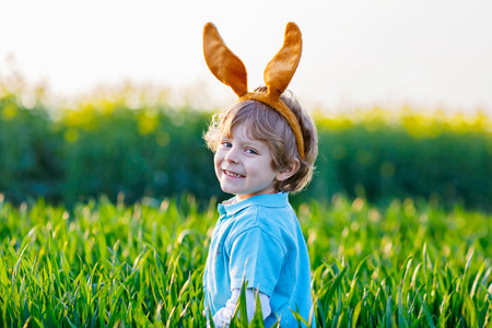 Cute little kid boy with bunny ears having fun with traditional Easter eggs hunt on warm sunny day, outdoors. Celebrating Easter holiday. Toddler finding, colorful eggs in green grass.の写真素材